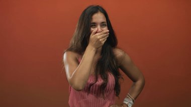 Ağzı kapalı ve gülümseyen pembe kolsuz bluz giyen genç bir kadın turuncu stüdyoda poz verdi..