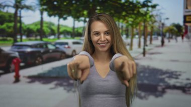 İki eliyle işaret eden ve arka planda arabalarla dolu güneşli bir şehir caddesinde gülümseyen bir kadın ve yürüyen insanlar, canlı bir şehir atmosferi yaratıyor..