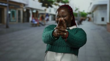 Yeşil kazaklı, kırmızı örgülü bir kadın ıssız bir caddede parmak tabancası jesti yapıyor. Kendine güveni ve oyunculuğu gösteriyor..