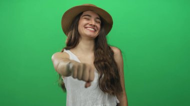 Beyaz atlet ve hasır şapka giyen genç bir kadın yeşil bir stüdyoda izleyiciye yumruk atıyor; dayanışma.