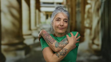 Gri saçlı son sınıf öğrencisi bir kadın, sokakta yeşil bir gömlek giyerek kendini kucaklıyor ve bulanık bir şehir geçmişine karşı dövmeler gösteriyor..