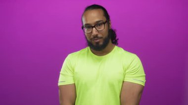Mavi stüdyoda gözlüklü ve görünür önkollu gülümseyen adam neon sarısı gömlek giyiyor..