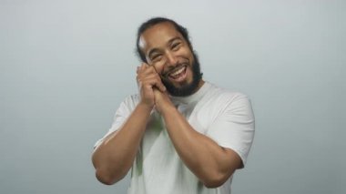 Soluk gri bir stüdyoda beyaz bir t-shirt giyerken çıplak kollarıyla el ele tutuşan, gülümseyen ve başını eğen bir adam; nazik bir sevinç.