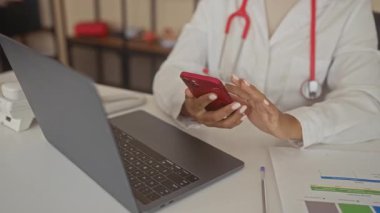 Kadın doktor, binadaki bilgisayarın yanında elleri ve steteskobu olan kırmızı akıllı telefonu dinliyor; odaklanın.