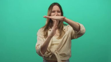 Young woman makes time out gesture with both hands near mouth and a pained expression in studio with teal green backdrop; disgust.