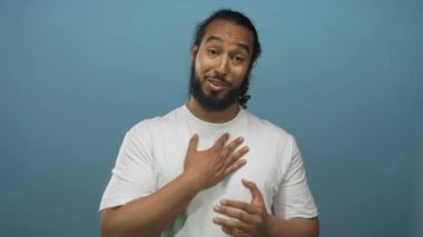 Mavi stüdyoda elleri göğsünün üzerinde gülümseyen beyaz tişört giyen ve minnettar görünen bir adam..