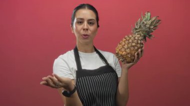 Çizgili önlük giyen esmer kadın pembe stüdyoda olgun ananas ve açık el hareketleriyle oyun meraklısı..