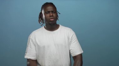 Çapraz kollu ve görünür kolları olan bir adam stüdyoda beyaz bir tişört giyiyor sert bir ifadeyle mavi bir arka plana karşı;.