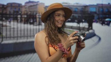 Bikini giymiş bir kadın elinde kamera, hareketli bir caddede duruyor, açık hava sahnelerini ilginç bir ifadeyle yakalıyor, modayı keşif ve gençlik ile harmanlıyor..