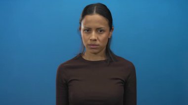 Latin kökenli genç bir kadın, izole edilmiş mavi bir arka plan duvarının önünde kulaklarını kapatır. Yüzünde bir ifade ve kahverengi gömlekle kollarını açar..