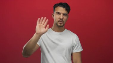 Genç adam canlı kırmızı arka plana karşı el sallıyor, günlük beyaz gömlek giyiyor, iletişim ve insan ilişkilerine odaklanmış dostça selamlaşma hareketi sergiliyor..