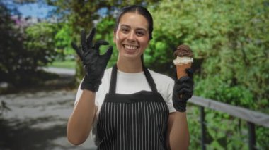 Önlüklü bir kadın dondurma külahı tutuyor ve ormanda kameraya gülümsüyor..
