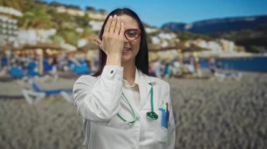 Steteskoplu beyaz üniformalı kadın doktor bir gözünü neşeli bir şekilde kapatıyor, güneşli bir plaj zeminine karşı ayakta duruyor, profesyonel ve sıradan atmosfer karışımını somutlaştırıyor..