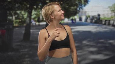 Kadın eli göğsünde, sonra sağ elini işaret ediyor. Spor sutyeniyle ve tozluklarıyla ağaçların ve binanın yakınındaki sokakta, çerçeveler boyunca gülümsüyor, minnettarlık..