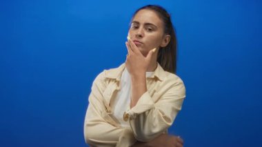 Mavi stüdyoda elleri çenesinde, sarı tırnaklı, bej gömlekli, kollarını kavuşturmuş ve düşünceli bir kadın..