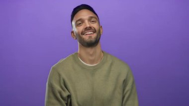 Genç İspanyol bir adam neşeli bir el hareketi yapıyor canlı mor bir arka plana karşı boynuzları andıran, arkadaşça ve eğlenceli bir şekilde gülümseyen.