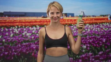 Spor sütyenli kadın iki eli göğsüne yakın yeşil meyve suyu şişesi tutuyor, gülümsüyor ve stüdyo lale sahasında poz veriyor;.