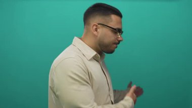 Gözlüklü genç İspanyol adam, canlı yeşil arka plan altında stüdyoda kameraya doğru elini uzatıyor; güven daveti.