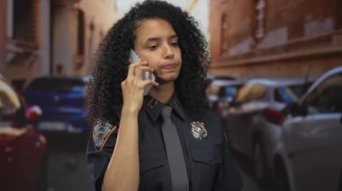 Üniformalı kadın polis memuru, park halindeki arabalarla ve şehir mimarisiyle dolu kalabalık bir caddede telefonla konuşuyor..