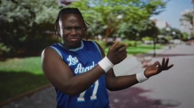 Mavi basketbol formalı adam 77 parmak çimdikliyor ve bilekliklerle sokağın kenarında park yolu ve ağaçların yanında asılıyor..