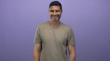 Stüdyoda gülümseyen ve kameraya işaret eden adam mor arkaplanlı bej tişört giyiyor..