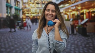 Elinde mavi bir cep telefonu olan genç bir kadın ve parke taşlı bir caddede kafenin ışıklarıyla izleyiciyi işaret ediyor; neşeli bir bağlantı.