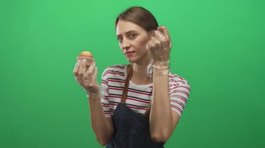 Parlak yeşil bir stüdyoda kot önlük giymiş, elinde temiz plastik eldivenli bir kek tutan genç bir kadın..