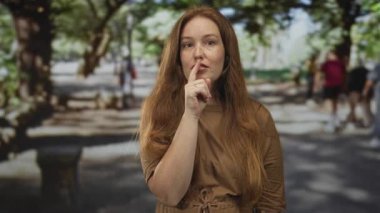 Kızıl saçlı genç kadın sessizlik için dudaklara parmak, güneşli yeşil parkta sakin bir yalnızlık..
