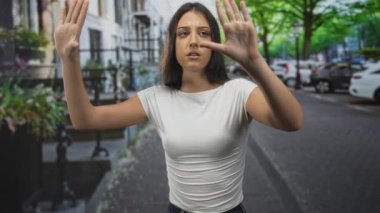 Kadın, şehrin bir caddesinde, beyaz tişört ve kot pantolon giymiş, avuçlarını ileri doğru uzatarak iki elini de kaldırır..