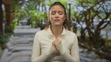 Parktaki İspanyol kadın duygusal görünüyor. Arka planda yeşil yapraklar var. Açık renk ceket giyiyor. Üzüntüsünü dile getiriyor..