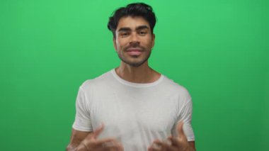 Latin asıllı genç adam yeşil stüdyoda avuç içi kaldırıyor beyaz tişört giyiyor ve hafif ışıklandırma altında belirsizlik var..