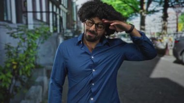 Kıvırcık saçlı bir adam saçlara dokunur, gözlük takar ve kaldırım kenarında mavi bir gömlek giyerken gülümser..