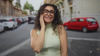 Gözlüklü ve kıvırcık saçlı bir kadın park halindeki arabalarla elini göğsüne koyup gülümsüyor..