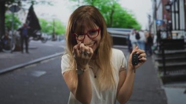 Kadın araba anahtarı tutuyor ve sokakta parmaklarıyla işaret ediyor, gülümsüyor ve kırmızı gözlük takıyor ve beyaz gömlek giyiyor; muzip.