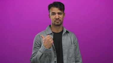 Gri ceketli genç ispanyol adam parlak pembe arka planda başparmak uçlarını işaret ediyor, kararsızlık ya da sorguyu kendine güvenen bir ifadeyle gösteriyor..
