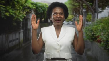 Avuç içlerini açık gösteren, yeşil çitli dar bir sokakta beyaz yelek giyen ve gülümseyen bir kadın..