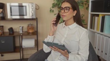 Ofiste elinde tablet ve telefon tutan kadın, sandalyede otururken gülümseyen ve gözlük takan kadın; uzaktan çalışma verimliliği.