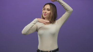 Mor bir stüdyoda elleri çerçeveli gülümseyen bir kadın, avuçlar başının yanında ve kol hareketi yükseltilmiş; sevinç güveni.