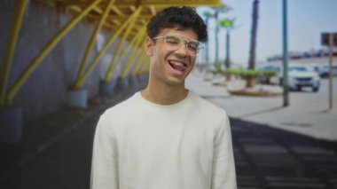 Gözlüklü bir adam, güneş ışığıyla parıldayan şehir sokağında dille göz kırpıyor. Gençlik enerjisi neşe saçıyor..