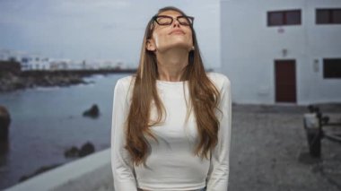 Gözlüklü kadın başını arkaya eğiyor ve deniz kenarındaki bir binada kapalı gözlerle gülümsüyor; sakin yansıma.