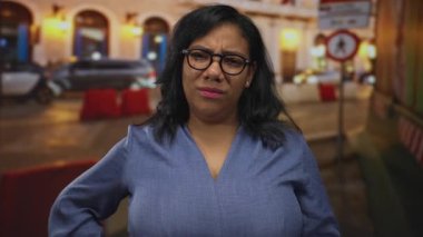 Gözlük takan ve mavi bluz giyen bir kadın gecenin bir yarısı elleri şehir caddesinde, arkasında bulanık araba ışıkları olan ellerle ayakta duruyor..