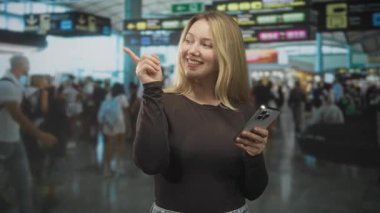 Uluslararası havaalanı terminalinde akıllı telefonu tutarken sol eliyle işaret eden gülümseyen sarışın kadın; neşeli seyahat özgüveni.