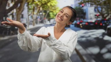 Elleri açık gülümseyen bir kadın beyaz bir bluz ve altın küpeler giyerek şehir caddesinde kameraya jest yaparak; dostça hoş geldin mutluluğu.