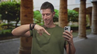 Adam akıllı telefonu sağ elinde tutarken parmağıyla işaret ediyor, eski kasabadaki sokakta yeşil gömlek ve bilezikler takıyor; dijital keşif merakı.