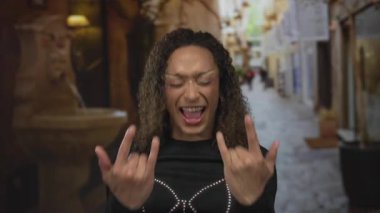 Kadın, tarihi binalarla dolu kalabalık dar bir sokakta elleri ile kaya boynuzları hareketi yapıyor; neşe gururu.