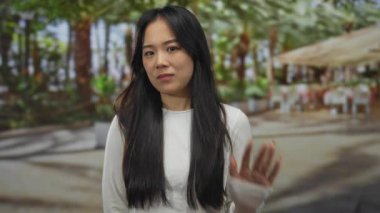 Beyaz tişörtlü, uzun saçlı genç bir kadın. Sokakta palmiye ağaçları ve masalarla dikiliyor. Huzurlu bir şehir anını yakalıyor..