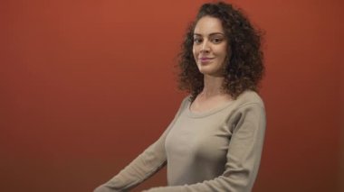 Turuncu bir stüdyoda bej rengi bluz ve kıvırcık saçlar giyen, hafif bir gülümsemesi olan, kendine güvenen bir kadın..