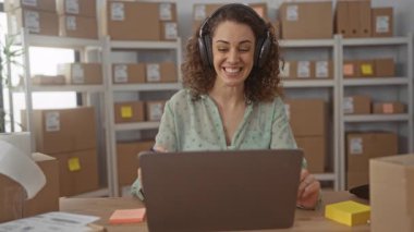 Kulaklık takan, gülümseyen ve dizüstü bilgisayarında kalemle işaret eden bir kadın, binada raflarda duran paketlerin arasında, küçük işletme destek sevinci.