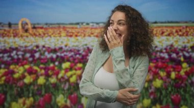 Renkli çiçeklerden oluşan stüdyo lale tarlasında elleri ağızda gülen genç İspanyol kadın, kolları gövde boyunca kıvrılmış; neşe pınarı kahkahası.