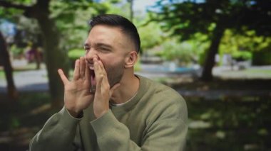 Çevresi canlı doğa ve ağaçlarla çevrili yeşil bir park ortamında bağıran genç adam dinamik ve canlı bir yüz ifadesiyle heyecanını dile getiriyor..